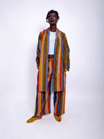 Load image into Gallery viewer, A person wearing a bold, multicolored striped Kente fabric outfit, consisting of a long coat and pants, against a white background.