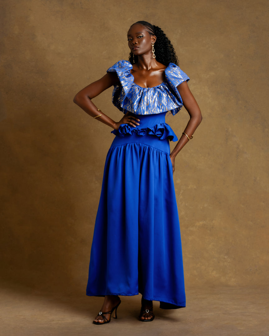 Woman wearing a blue outfit with ruffled top and wide-leg pants against a brown background